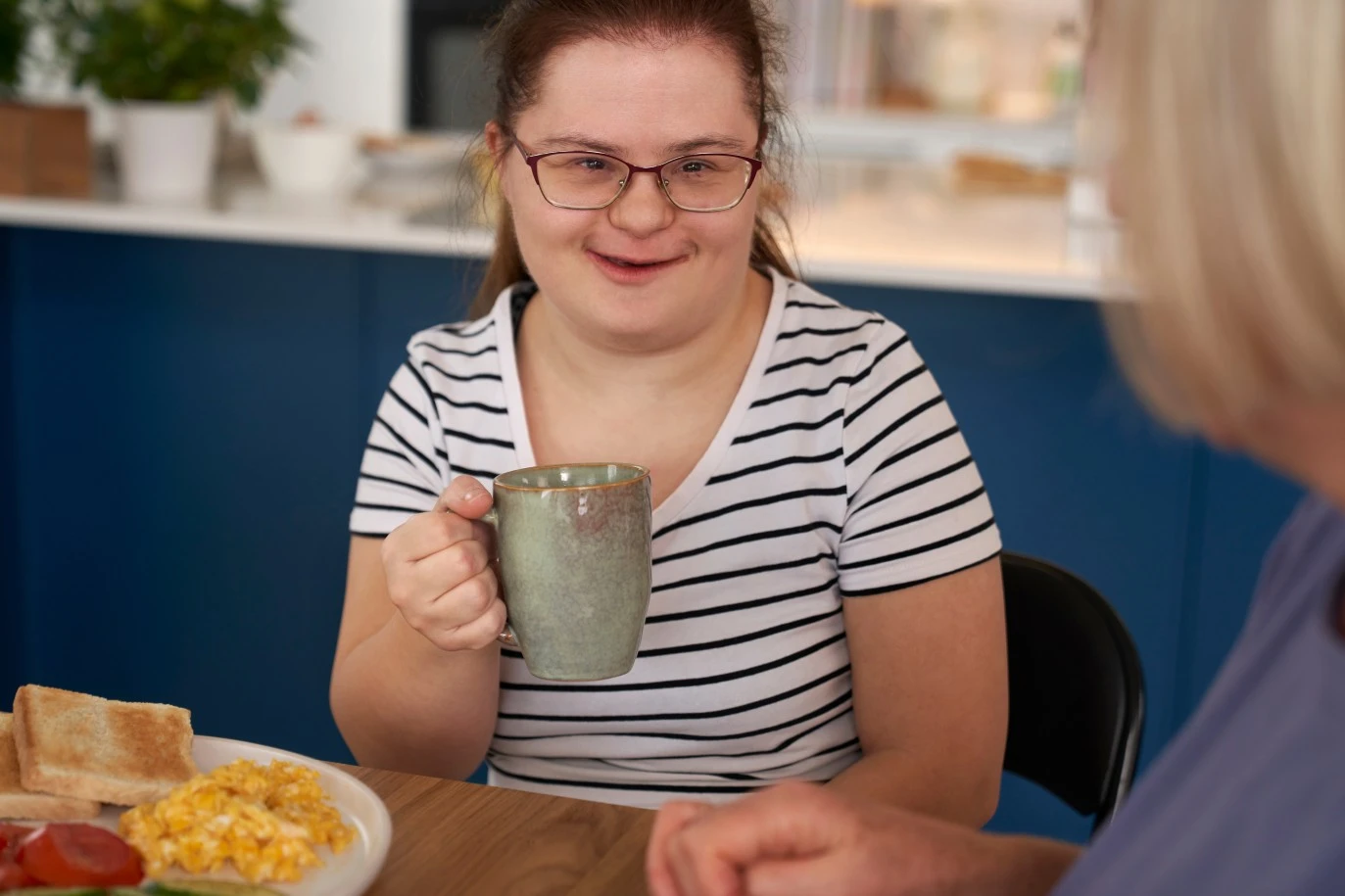 Disabled girl with down syndrome holding mug while talking to support worker at kitchen table