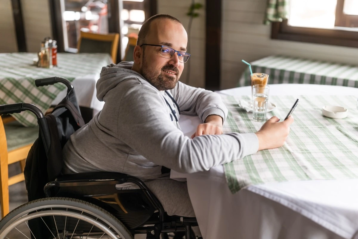 Disabled paraplegic man in wheelchair sitting at cafe table and smiling at camera while holding phone